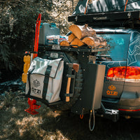 Off-road SUV with rear cargo rack carrying firewood, gear bag, and tools in a forest setting